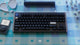 DSS Sad Girl keycaps render. Designed by Mintlodica. Black mechanical keyboard with silver edges with dark keycaps. The keycaps have glowing blue alpha texts. iPod, broken heart tray, matching switches on a picnic patterned deskmat.